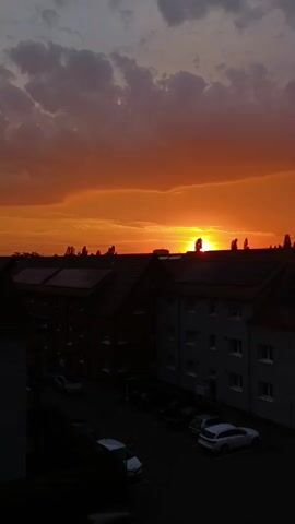 The video shows a sunset over a residential area. The sky is filled with vibrant orange and red hues, with some dark clouds scattered throughout. In the foreground, there are buildings with dark roofs, some of which appear to have solar panels. There are also cars parked in a lot below the buildings, mostly obscured by shadows. The sun is setting behind a line of trees, casting a silhouette against the bright sky. The overall impression is a peaceful, serene evening scene.
