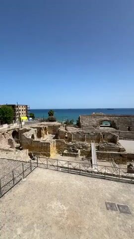 The video shows a wide shot of an ancient Roman amphitheater under a clear blue sky. The amphitheater is made of stone and is partially ruined, with sections of seating and walls still intact. The arena floor is visible in the center, and a wooden ramp leads up to one of the lower levels. Beyond the amphitheater, the blue ocean stretches to the horizon, with several ships visible in the distance. In the background to the left, there is a modern yellow building. To the right, there are some palm trees and greenery on a hill overlooking the amphitheater. The foreground of the video is a sandy, dirt area with a metal fence running along the edge of the amphitheater. There are no people visible in the scene. The camera is stationary.