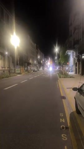 The video shows a nighttime view of a street with buildings on either side. Streetlights illuminate the road, and car headlights are visible in the distance. The road has white lane markings, and there are yellow markings and lettering on the right side of the road. There are some trees and planters along the sidewalk. The buildings appear to be multi-story with windows. The camera is moving, giving a sense of walking down the street. There are no visible people in the foreground.