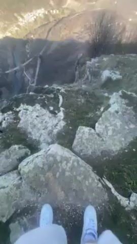 The video shows a scenic view from the top of a rocky cliff on a sunny day. Initially, the camera focuses on the person's feet wearing white sneakers and a portion of their white pants, then pans upward to reveal the expansive view of a valley. The landscape includes winding roads, a town nestled in the valley, and mountains in the distance, all under a clear blue sky with a few scattered clouds. The viewpoint is partially covered with rocks and sparse vegetation, creating a natural foreground to the panoramic vista.