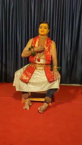 A male performer, dressed in traditional Indian attire for a dance performance, sits on a stool against a dark curtain backdrop. He has elaborate yellow face paint with red accents around his eyes and mouth, a bright red vest, and a golden belt. His legs are bent, and his feet are bare, adorned with ankle bells. He wears armbands with green pom-poms and a golden necklace. The stage floor is covered in red carpet. The video shows him in a static pose, likely at the beginning or end of a performance.