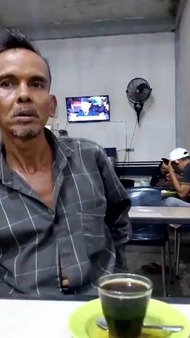 The video shows a man in a gray plaid shirt sitting at a table with a glass of dark liquid on a green saucer in front of him. He is looking slightly off-camera and talking. In the background, there is a television playing news, a black fan, and two other people at a table. One person is wearing a white cap and is taking a photo or video with a smartphone. The environment appears to be a simple, possibly public eating area.