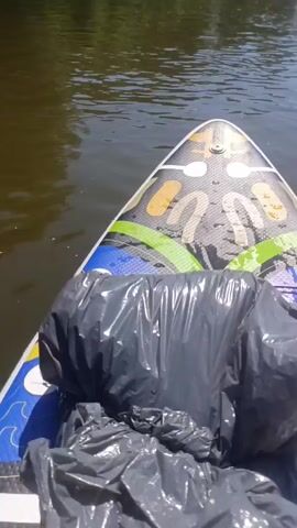The video begins with a first-person perspective of someone on a paddleboard on a river. The paddleboard is colorful with a black plastic bag on the front. The river is calm and murky brown, with green trees and vegetation on the banks. The sky is clear and blue.

The camera then pans to show other people on paddleboards further down the river. There are at least four other people visible, all on paddleboards. One person, wearing a light-colored top, is seated on their paddleboard, holding a paddle. Another person, wearing a white cap and white top, is also seated and paddling. Two other individuals are further in the distance, also on paddleboards. The overall environment is a calm, sunny day on a river surrounded by lush greenery.