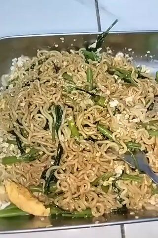 The video shows a close-up, overhead shot of a metal tray filled with instant noodles mixed with green vegetables and scrambled eggs. A fork is seen stirring the noodles, lifting some of them to reveal the texture and ingredients. The noodles appear to be seasoned and lightly browned. The background is blurred but suggests a kitchen environment with white tiled surfaces. The lighting is bright, illuminating the food clearly.