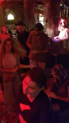 The video shows a group of people dancing in a dimly lit room, likely a bar or club, illuminated by red lighting. The floor is covered with an oriental rug. A woman in a tube top and loose pants dances barefoot near the center of the frame, with other people dancing around her.