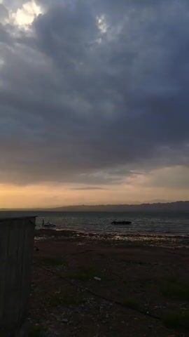 A vertical video captures a wide view of a body of water under a cloudy sky. The water appears choppy and a dark boat is visible on the water in the middle ground. The shore in the foreground is composed of dark sand and gravel with some sparse green vegetation. In the distance, a range of low, dark mountains is visible under a hazy, pale orange horizon. The sky is filled with heavy, dark grey clouds, with a sliver of lighter sky visible near the horizon. The scene appears to be either dawn or dusk. The video begins with a close-up of a wooden post or structure on the left side of the frame and pans slowly to the right, revealing more of the shoreline and the water. There are no people visible in the video.