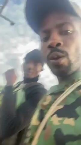 The video shows two men in camouflage clothing walking on a dirt road, possibly in a rural area. The man in the foreground is wearing a camouflage shirt and a baseball cap. He is holding the camera and speaking. The man in the background is wearing a black turtleneck sweater, a black beanie, and camouflage pants. Both men appear to be carrying rifles or similar long firearms over their shoulders. The environment appears to be a village or rural area with green vegetation and simple buildings in the background. The weather appears overcast.