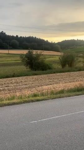 The video shows a rural landscape at sunset. A paved road runs along the bottom of the frame. Beyond the road, a harvested field, likely for grain, is visible with stubble remaining. Further back, there are trees and rolling hills covered in vegetation. The sky is filled with clouds, and the setting sun casts a warm, golden light, creating a hazy glow. Power lines and a utility pole are also visible in the distance. The camera appears to be stationary, capturing a panoramic view of the natural scenery.