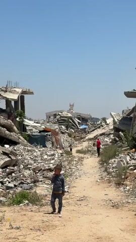 The video displays a bleak landscape of destruction in Jabalia, Palestinian Territories. The scene is dominated by the remnants of bombed-out buildings, with piles of rubble and twisted metal filling the frame. The sky is a clear, bright blue, suggesting a sunny day, likely midday given the harsh shadows.

In the foreground, a young child, dressed in a grey tracksuit, stands on a dirt path and looks towards the camera with a concerned expression. Another child, further down the path, is seen walking away. In the mid-ground, a person in a red shirt is also walking along the path, their back to the camera. Towards the left of the frame, a man in a light-colored shirt and dark pants walks into the scene.

The background is filled with more destroyed structures, some with rebar exposed, indicating significant damage. Faded tarps are visible stretched over some of the debris, possibly providing makeshift shelter. The overall atmosphere is one of devastation and hardship, with the presence of children amidst the ruins evoking a sense of loss and resilience. There is no visible text in the media file.