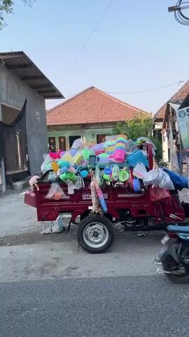 The video begins with a stationary, dark red pickup truck laden with an assortment of colorful plastic goods, including buckets, containers, and toys. The truck is parked on the side of a paved road in what appears to be a residential or commercial area. In the background, there are buildings with tiled roofs and some smaller structures. The sky is clear and bright, suggesting daytime.

The scene then shifts to a different perspective, focusing on a roadside stall with blue and yellow signage. The stall appears to be selling drinks, with an advertisement featuring ice tea. A black motorcycle with a rider wearing a dark jacket and helmet is seen passing by in the foreground, moving from right to left across the frame. The rider is partially obscured by the movement of the motorcycle. The environment seems to be a street with buildings and vegetation visible. The overall impression is a glimpse of everyday life in a street setting, possibly in Southeast Asia, given the visual cues.