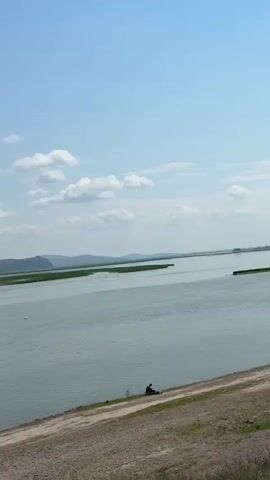 The video shows a scenic view of a lake with rolling hills in the background under a clear blue sky with scattered clouds. In the foreground, there is a dirt path and grassy shoreline. There are a few people visible on the shore, one appearing to be fishing, and a car is parked on the side of the path. The water is calm and reflects the sky and landscape.