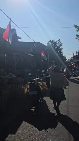 A street scene under a bright sun. Two Indonesian flags, red and white, are visible. Several people are present, some walking and some on motorcycles. A motorcycle is parked in the foreground, loaded with yellow bags. A person is walking away from the camera down the street. Other people are seen further down the street, some appearing to be vendors with goods on display, including what looks like produce. The environment appears to be a residential or market area with simple buildings lining the street.