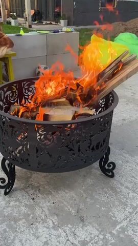 Here is a description of the video:

The video shows a metal fire pit with a roaring fire in a backyard. Someone is feeding the fire with wood scraps.  The pit is surrounded by a concrete patio. In the background are piles of dirt, a partially constructed building covered in black wrap, a finished home, yellow and green beanbag chairs, and a swimming pool. A woman in a light green tracksuit walks in front of the camera, and then raises her hands to her face.