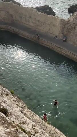 The video shows a view looking down into a natural swimming pool carved into a rocky coastline. The water is clear and turquoise, with sunlight reflecting off its surface. Two people are swimming in the pool, one in a dark swimsuit and the other in a red swimsuit. To the left of the pool, a steep, rocky incline covered in sparse vegetation slopes down towards the water. On the right side of the pool, a stone wall forms the edge, with a metal ladder leading down into the water. A few items, possibly bags or towels, are placed on the walkway next to the wall. In the distance, the open sea is visible, with waves breaking against dark rocks. The scene appears to be set during the daytime, likely late morning or early afternoon given the bright sunlight. The setting suggests a coastal resort or a popular swimming spot. The overall atmosphere seems relaxed and summery. There is no visible text in the media file.