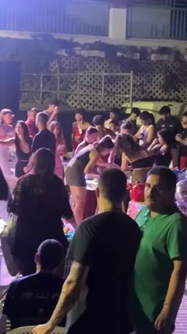 The video shows a crowded outdoor gathering at night. A group of people are standing around tables, some appearing to be serving food or drinks. Others are milling about, some in conversation, others dancing. The lighting is dim and appears to be from artificial sources, casting a purplish hue on some of the attendees and the surrounding environment. There are plants and vines visible in the background, suggesting an outdoor patio or garden setting. The overall atmosphere appears to be social and lively, possibly a party or event.