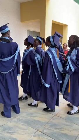 A group of students in blue graduation gowns and caps are standing in a line, facing away from the camera. They appear to be in a hallway or outdoor area with tiled floors and building structures visible in the background. The students are facing towards a doorway or entrance. One student in the foreground is looking back towards the camera. The students are holding their gowns and adjusting their caps. There are other people, possibly staff or other students, in the background. The overall atmosphere appears to be one of anticipation or preparation for an event.