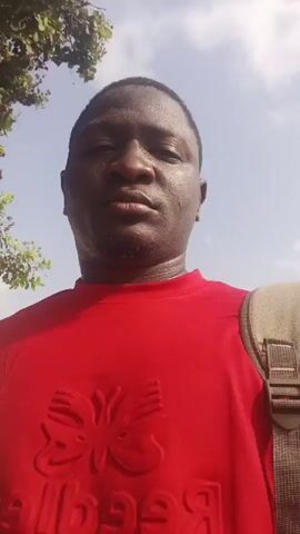 The video shows a man in a red shirt with a butterfly logo, standing outdoors on a sunny day.  He appears to be speaking to the camera. There's a leafy tree behind him on the left, and a glimpse of the sky above.  A strap is visible on his right shoulder, suggesting he may be carrying a bag. The camera is positioned at a low angle, looking up at the man.