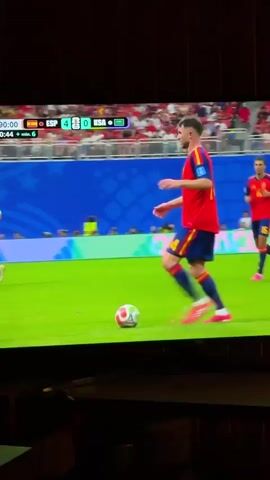 The video shows a soccer match on a large screen television. The scoreboard indicates the score is ESP 4, RSA 0, and the game is in the 90th minute. A player in a red and yellow uniform, wearing number 14 and the name "LAPORTE" on his back, is controlling the ball with his feet. He is on a bright green soccer field, with other players in white and red uniforms in the background. There are spectators in the stands, some wearing red. The scene is viewed from a slightly elevated angle, with the bottom of the television screen and a dark foreground area visible.