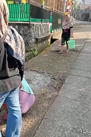 The video shows an outdoor scene on a street, with a group of people. In the foreground, a person wearing a black shirt and blue jeans is partially visible, facing away from the camera and appearing to be interacting with someone or something on the ground. To their left and slightly behind, another person wearing a light blue top and purple pants is crouched or kneeling. In the middle ground, a person wearing a hijab and a dark t-shirt with a graphic, along with dark shorts, is walking towards the right, carrying a green bucket in their right hand. They are walking on a concrete path next to a shallow, dirt-lined ditch. Behind the walking person, a fence made of wooden poles and black metal is visible along a wall. The environment appears to be a rural or suburban street with some vegetation and buildings in the background. The lighting suggests it is daytime. The dialogue in the video is in Indonesian and translates to "This year I'll trade it for tempeh, Sir. Yes." and "I'll trade this year for tempeh, Sir."