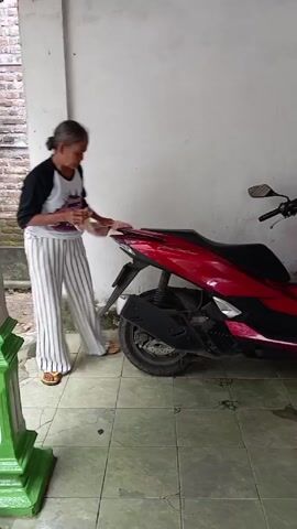 Here is a concise description of the video:

The video shows a person cleaning a red scooter with a cloth, near a white wall and a tile floor.  The person is wearing striped pants and a shirt with dark sleeves and a white front. Another person is sitting on a small wooden bench nearby, watching the cleaning. A wooden door is visible in the background.
