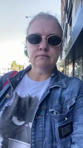 A woman is recording a selfie video outdoors. She has blonde hair pulled back in a bun, is wearing sunglasses, a white t-shirt with a cat graphic, and a denim jacket. She makes a peace sign with her hand. The background includes a street, buildings, and trees. The video appears to be shot during the day with good lighting.