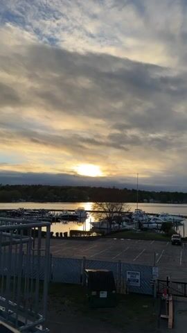The video shows a sunset over a harbor filled with boats. The sky is partly cloudy, with the sun peeking through the clouds. The sun's reflection is visible on the water. In the foreground, there is a parking lot with a dumpster and a sign that says "Members Only." A metal railing is visible in the lower left corner.