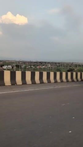 A first-person perspective video shows a person riding a scooter on a highway. A concrete barrier with black and white stripes lines the side of the road. In the distance, power lines and buildings are visible. The sky is overcast with scattered clouds. The rider's hands, wearing a watch, are visible on the scooter's handlebars. The side mirror reflects the rider's face and shoulders. A truck with a flatbed trailer is partially visible in the foreground in the first few frames. The video continues to show the highway and the surrounding landscape.