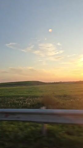 The video shows a view from a moving vehicle of a rural landscape at sunset. The sky is a gradient of blue and orange, with wispy clouds. A large, bright sun is visible on the horizon, casting a warm glow over the scene. Fields of green vegetation, possibly crops or wild grasses, stretch across the middle ground, with rolling hills in the distance. Trees and bushes line the side of the road, their silhouettes dark against the bright sky. A metal guardrail is visible in the foreground, indicating the perspective is from a road or highway. The overall impression is one of a peaceful and scenic sunset drive.