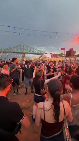 A crowd of young adults is gathered outdoors at what appears to be an event or festival. They are dancing and socializing. String lights are strung overhead, and there are buildings and a bridge in the background. The sky is overcast, suggesting it might be evening or a cloudy day. A DJ booth or stage with speakers and lighting is visible on the right side of the frame. The ground appears to be sandy or dirt, with some wet patches, possibly indicating recent rain. The overall atmosphere seems lively and festive.