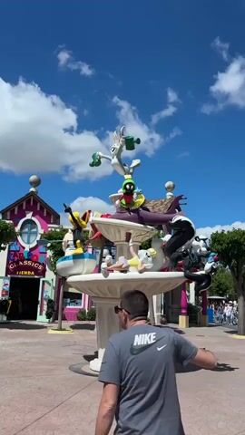 The video shows a plaza area in a park under a sunny sky with scattered clouds. A large, tiered fountain is centered in the plaza, adorned with statues of various Looney Tunes characters. Several people are walking around the plaza, including some passing by the fountain. The surrounding buildings have cartoonish, colorful designs with storefronts and marquees, and lush green trees are interspersed throughout the area.