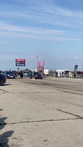 A video captures a drag race between a blue and a black Volkswagen Golf on an asphalt track, lined with spectators behind barriers under a partly cloudy sky. The cars accelerate quickly, generating smoke. In the background, banners and promotional material are visible. The camera remains stationary throughout the event.
