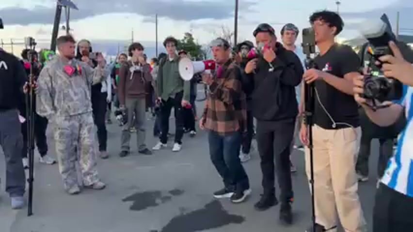 A group of approximately 15-20 individuals is gathered outdoors on a paved surface, likely a street or parking lot, under an overcast sky at dusk or early evening.

The central figure, a man in a plaid shirt, clear goggles, and pink respirator filters, is speaking passionately into a red and white megaphone, gesturing with his right hand. He is addressing the crowd, stating, "People have been saying that you have been going inside this building and assaulting these..."

Many of the attendees, predominantly young men, are also wearing respirators or gas masks with pink or black filters. Several individuals are actively filming the scene with smartphones, one of which is mounted on a tripod to the right of the speaker. Other subjects are observing, some holding cameras or wearing backpacks. The man on the far left wears a camouflage-patterned hoodie and pants, also with pink respirator filters.

The overall atmosphere suggests a protest or public gathering, with a focus on recording the event. Utility poles and power lines are visible in the background, hinting at an urban or industrial setting.