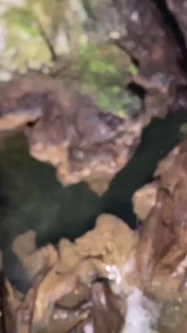 The video shows a close-up view of a small waterfall cascading into a pool of dark water within a cave. The cave walls are rough and irregular, with formations of brown and tan rock, and patches of green moss. The water is clear enough to see the bottom of the pool in some areas. The lighting is dim, suggesting an underground environment. There are no people or animals visible in the video. The focus is on the natural geological features and the flow of water.