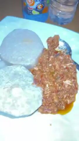 A top-down video shows a white plate with two circular flatbreads, likely pounded yam, beside a scoop of a reddish-orange stew. A spoon is in the stew. A bottle of “Active Zero” orange juice and a clear water bottle are behind the plate. The table surface is brown. The camera moves slightly, keeping the plate and food in view.