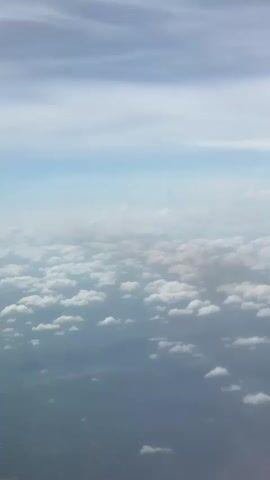 The video shows a view from above, likely taken from an airplane, looking down at a layer of puffy white clouds. Above the cloud layer, the sky is visible, with light blue transitioning to areas of thinner, higher altitude clouds.