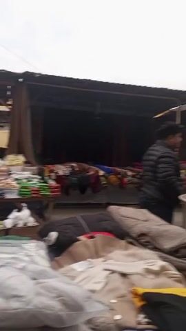 The video shows a bustling outdoor market on an overcast day. The camera pans slowly, revealing rows of stalls under metal roofs, laden with various goods. A large metal tandoor oven stands prominently in the foreground, with two men nearby. One is wearing a black puffer jacket and the other a blue jacket and black beanie. There are also stacks of plates on a small white table next to the tandoor oven.  In the foreground, there is a pile of clothes. In the background, there are many people milling about.