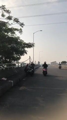 Two individuals are interacting with a parked motorcycle on a roadside. One person, wearing a red cap and a maroon shirt, is crouched down, appearing to work on the rear of the motorcycle. The other person, wearing a dark shirt and a baseball cap, stands beside the motorcycle, looking at it. A second motorcycle with a rider is visible further down the road, moving away from the camera. The scene takes place on an elevated road, possibly a bridge, with a metal railing and concrete barriers. Trees are visible on the left, and a hazy sky suggests it might be morning or late afternoon. Power lines are also present overhead.