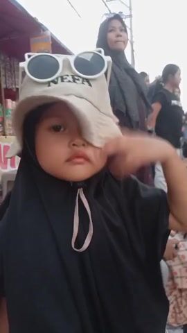 The video shows a young child in a bucket hat and sunglasses, wearing a black headscarf. The child is looking to the left and making a hand gesture. In the background, several other people are visible, some wearing headscarves. There appears to be a street market or outdoor event, with stalls and goods in the background. The lighting suggests it is daytime.