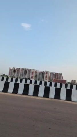 The video shows a view of a highway with a rider on a motorcycle in the foreground. In the background, there are several tall apartment buildings and a clear blue sky. The highway is separated by a barrier with black and white stripes. The rider on the motorcycle is wearing a helmet and is moving across the frame.