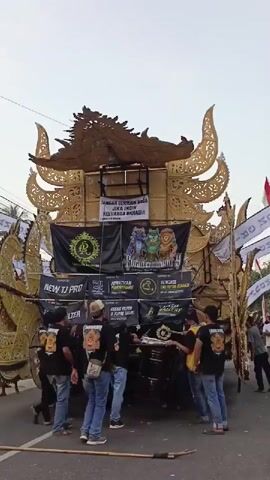 The video shows a group of young men, most of whom are wearing black t-shirts with a graphic of a lion's head and text that reads "NEW TJ PRO," gathered around a large, ornate float or vehicle. The float is decorated with golden, elaborate carvings, resembling mythological creatures or traditional motifs. It is adorned with several banners, predominantly black with yellow and white lettering, and features large speakers stacked on top. Some banners display logos with a stylized "R" and other graphics. The men appear to be interacting with the float, possibly adjusting equipment or preparing for an event. They are standing on a paved road, and in the background, there are hints of a crowd or other people, as well as some white banners with text, suggesting a public gathering or parade. The overall impression is that of a mobile sound system or performance stage being prepared for a celebration or event. The lighting suggests it is daytime, possibly late afternoon given the sky's hue.