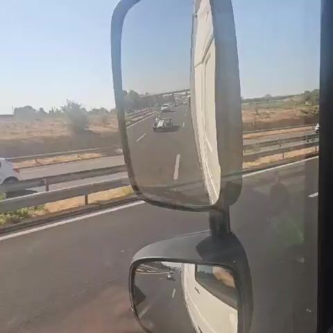 The video shows a side-view mirror of a truck. Through the mirror, a blue sports car with white stripes is visible driving on a highway. The car is in front of the truck. Other vehicles and a bridge are also visible in the distance. The sky is clear and blue, and the landscape appears dry and sunny. The video is shot from the perspective of the truck driver.