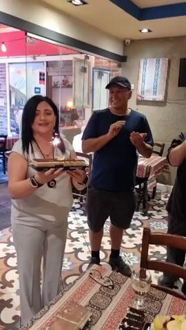 A woman stands in what appears to be a restaurant holding a cake with sparklers lit on it. She is wearing a light gray top and pants, and a necklace with a dark pendant. She is blowing on the sparklers. Behind her, a man wearing a blue t-shirt and a baseball cap is clapping. The restaurant has tiled floors with a patterned design and tables with patterned tablecloths. There are other cakes on the table in front of the woman. The background shows a storefront with windows and red awnings. The video is short, capturing a brief moment of celebration.