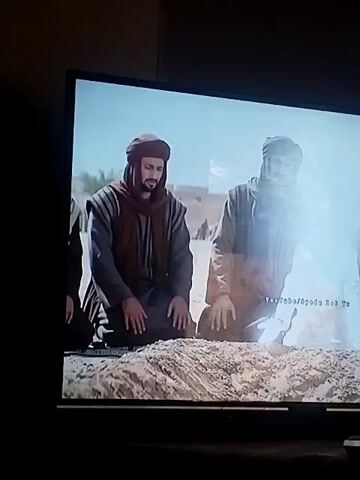 Three men are kneeling on sand, facing the camera. The man on the left is dressed in dark robes with a brown scarf. The man to his right is in lighter robes with a dark head covering. Between them, closer to the camera, is a fourth figure in white robes, barely visible. They appear to be outdoors, in what looks like a desert or sandy environment. In the background, there are building structures suggesting a city or settlement. The scene is likely from a historical or religious film or TV show. A YouTube watermark is in the bottom right corner.