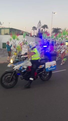 The video shows a procession on a street with palm trees. The procession consists of women in silver costumes with colorful feathers and headpieces. A police officer on a motorcycle leads the procession. Spectators line the side of the street to watch.