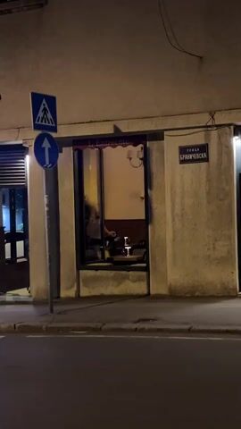The video shows a street view at night in front of a building with a store or restaurant. There is a sidewalk and a street in the foreground. A street sign with Cyrillic writing is visible on the building. A pedestrian crossing sign and a directional arrow sign are on a pole to the left. The building has large windows and an open doorway. Inside, there are tables and chairs. A poster with a woman in a red dress is hanging on the wall inside. The lighting is warm, suggesting interior lights are on. The overall scene is calm and depicts an urban environment at night.