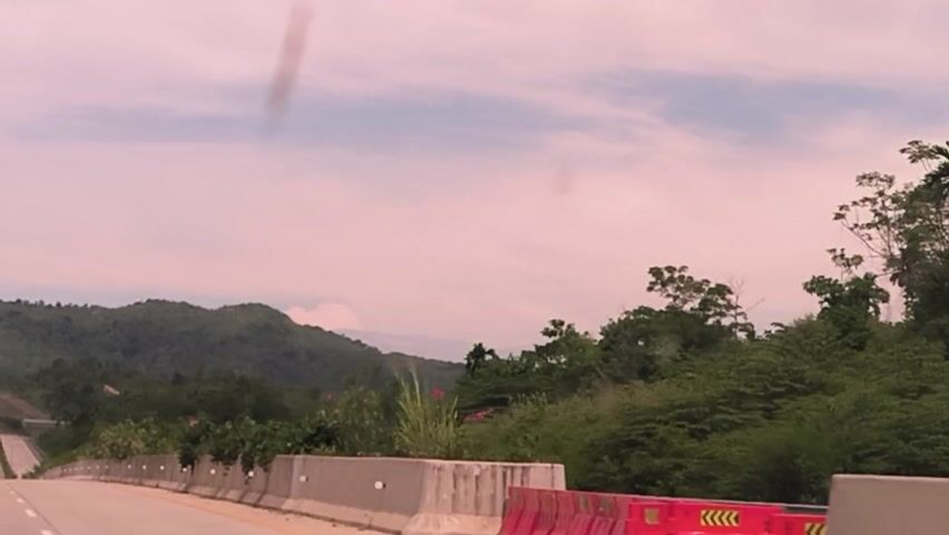 The video depicts a view from a vehicle moving along a highway with surrounding lush green vegetation. The camera captures the highway, adjacent green hills, and the sky. The sky is a muted, somewhat pinkish tone, likely due to atmospheric conditions or a filter. A vehicle can be seen traveling ahead on the highway. The camera angle is slightly tilted.