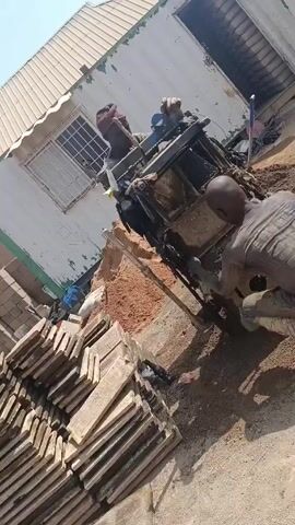 Two men are working at an outdoor construction site on a sunny day. One man is seated operating a machine, possibly a concrete mixer, which is placed on a small mound of reddish-brown dirt. The other man is kneeling on the ground next to the machine, holding what appears to be a long wooden tool or lever. There is a stack of rectangular concrete blocks in the foreground and a small white building with a metal roof and barred window in the background. Construction debris litters the ground.