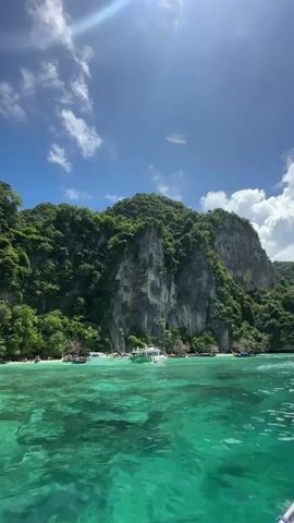 The video shows a body of water with boats. A large, rocky island covered in lush green trees rises behind the water. The sky above is blue with scattered white clouds. The water appears clear and turquoise. Several boats are visible in the water, some near the shore of the island. The sun is shining, casting light and shadows on the scene.