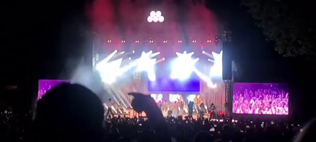 A nighttime outdoor concert is underway. A large stage is illuminated by bright spotlights and a backdrop of lights. A band is performing on stage, with singers and musicians visible. Large screens on either side of the stage display images of the performers. A crowd of people is gathered in front of the stage, many holding up their phones to record the event. Smoke or haze is visible on stage, adding to the atmosphere. The overall scene suggests a lively and energetic performance with an engaged audience.