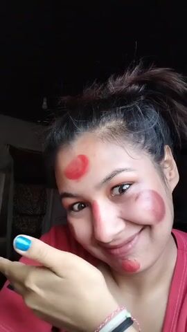 A young woman with fair skin is the subject of this close-up video. She has dark hair pulled up in a messy bun, and her nails are painted a bright blue. She's wearing a red t-shirt.  The most striking feature is the makeup she is wearing, which consists of a red circle on her forehead, a red line down the bridge of her nose, and brown circles on her cheeks. She smiles at the camera and playfully covers her face with her hand. Her eyes cross slightly as she covers her mouth and nose. The background is dark, possibly a room with a shadowed doorway or window behind her.